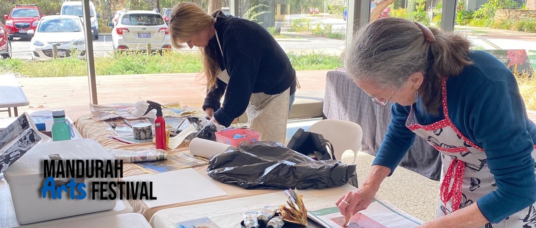 Two women are bent over tables covered with paper, and art supplies, working intently on their art pieces